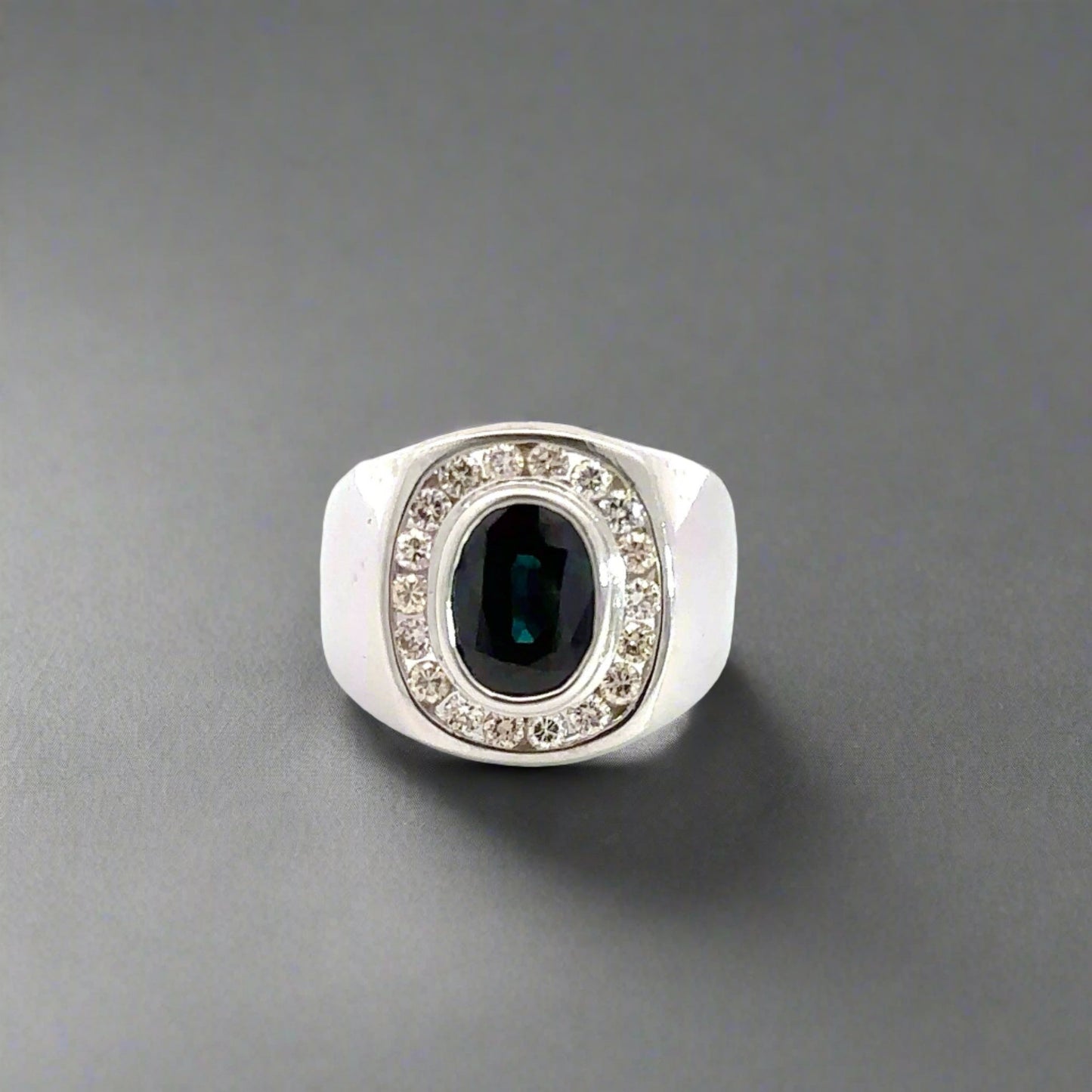 Front of white gold dome shaped ring with 1 dark blue oval-shaped Sapphire in center and 18 round diamonds surrounding it. Shows scratches on gold