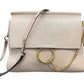 A light beige leather shoulder bag with a flap closure, a large gold-tone O-ring, a gold chain detail, and a thin black-and-beige strap, shown on a plain white background.