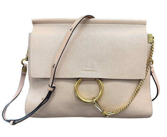 A light beige leather shoulder bag with a flap closure, a large gold-tone O-ring, a gold chain detail, and a thin black-and-beige strap, shown on a plain white background.