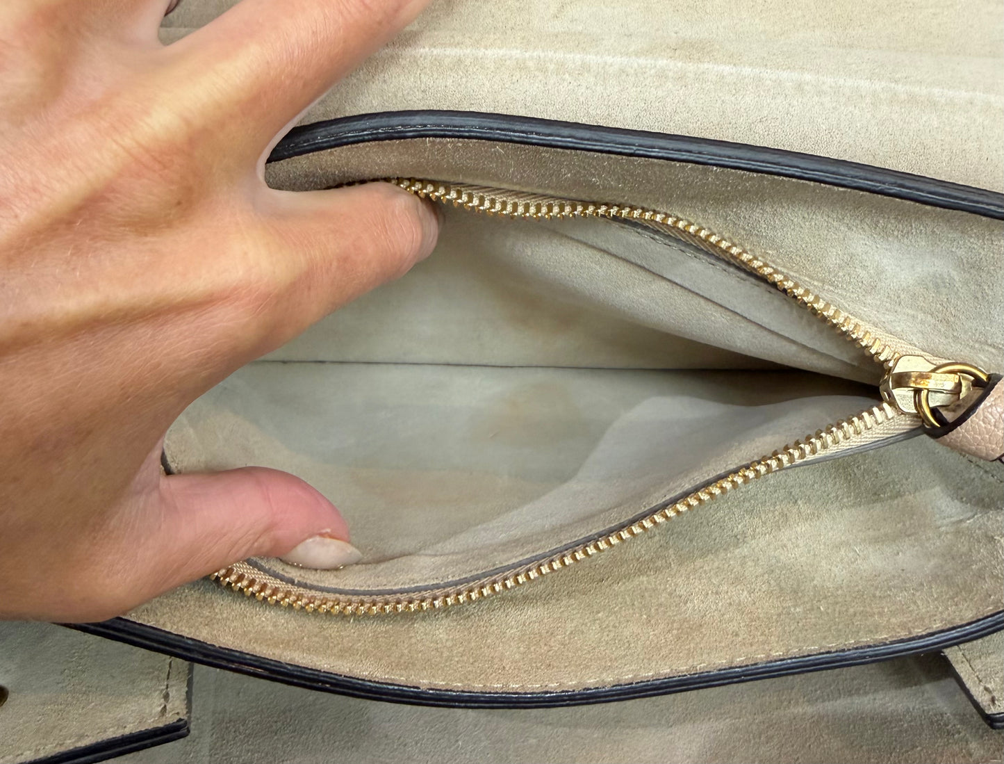 Close-up of a hand opening the interior beige suede zipper pocket with a gold zipper and black trim.