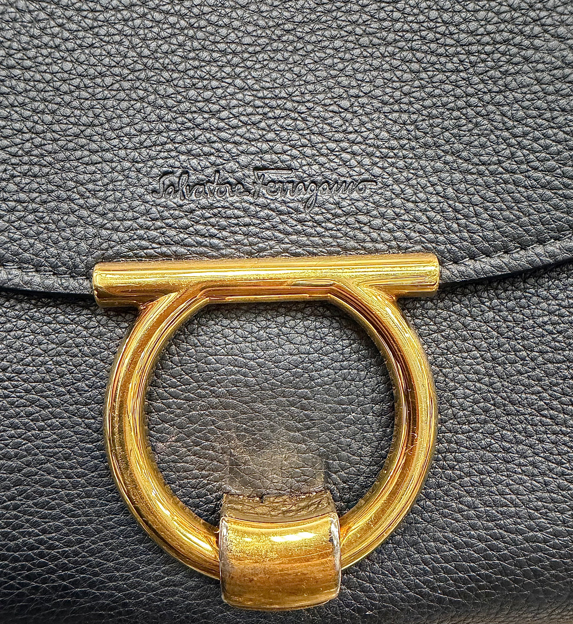 Close-up of a black pebbled leather handbag featuring a large gold-tone metal O-ring clasp and an embossed "Salvatore Ferragamo" logo on the flap.