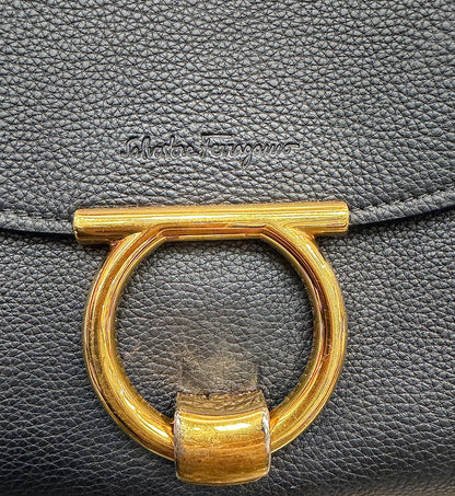 Close-up of a black pebbled leather handbag featuring a large gold-tone metal O-ring clasp and an embossed "Salvatore Ferragamo" logo on the flap.