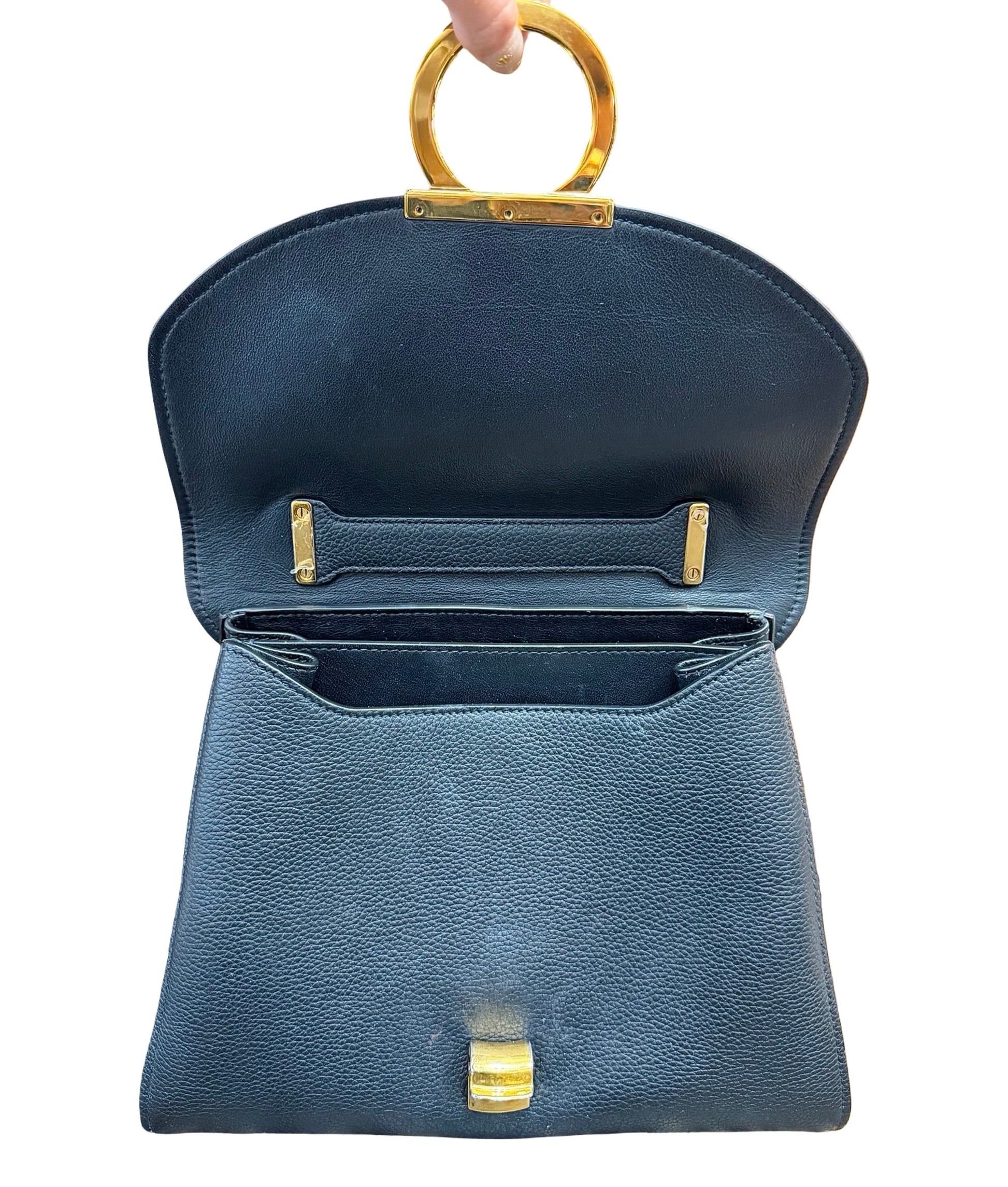 A black pebbled leather handbag with gold-tone hardware, shown open with a circular top handle being held by a hand against a white background.