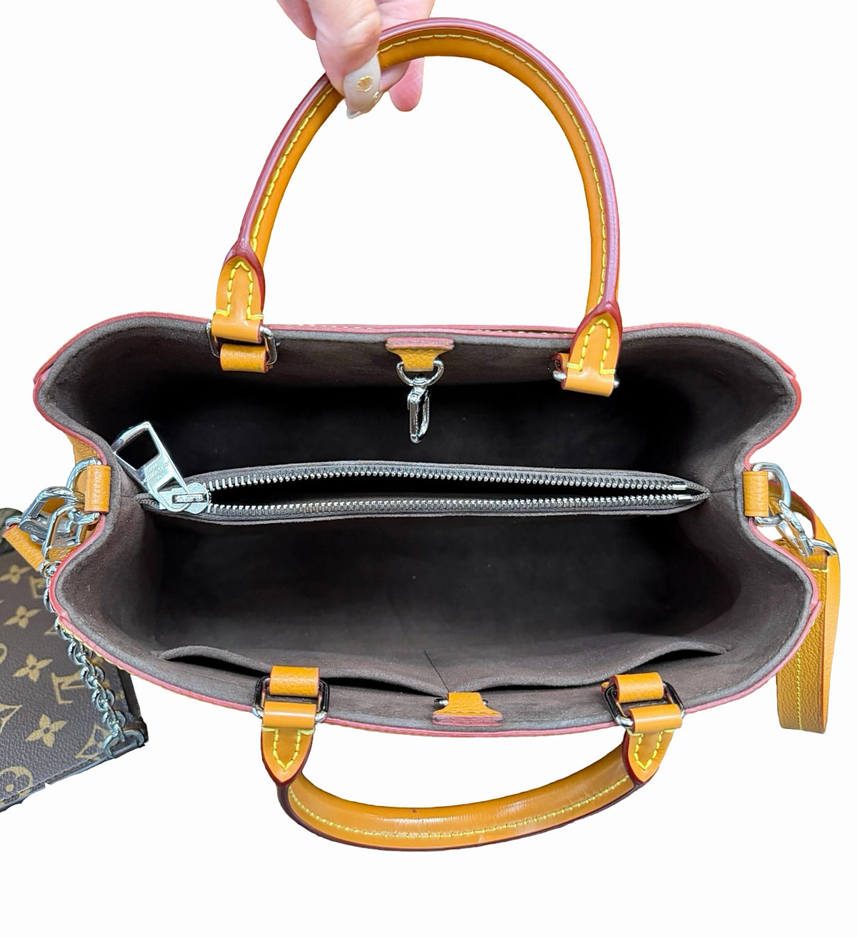 An open Louis Vuitton handbag shown from the inside, with a hand holding the top handle. The bag has brown leather handles and trim, a dark brown interior, and silver-tone hardware.