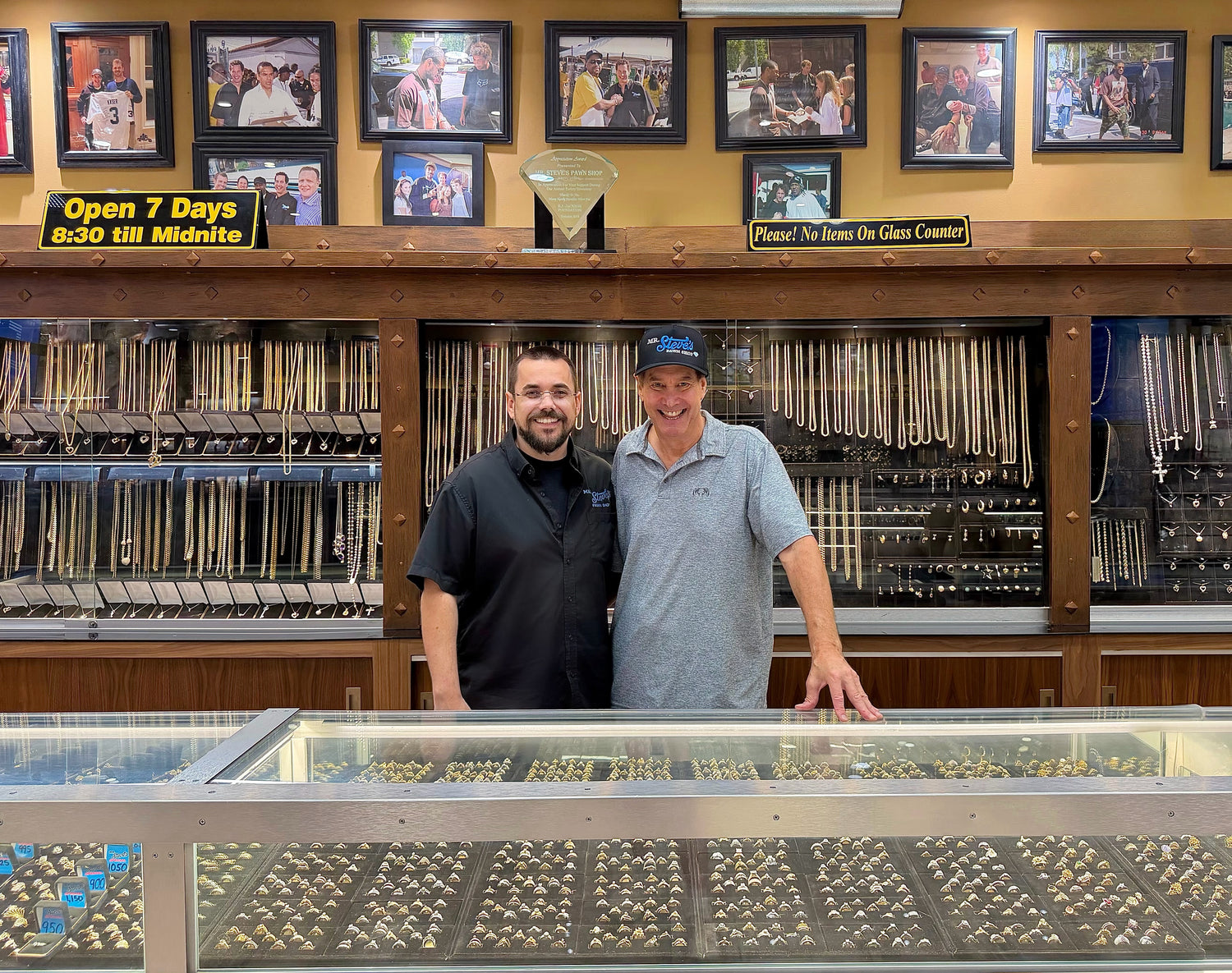 Two men standing behind a counter in a store with shelves and signs in the background.